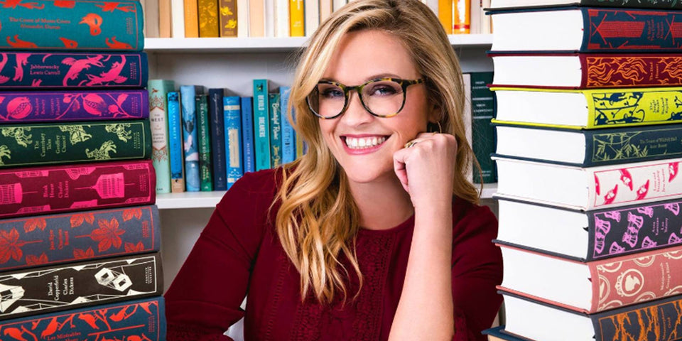 Reese Witherspoon smiling between tall stacks of books.