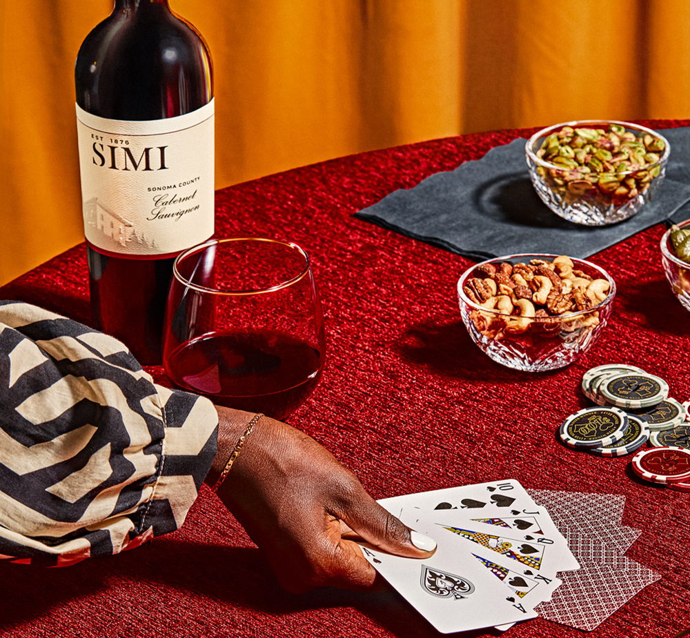 A bottle of SIMI Cabernet sits on a table, a glass of wine and a woman's hand holding playing cards.