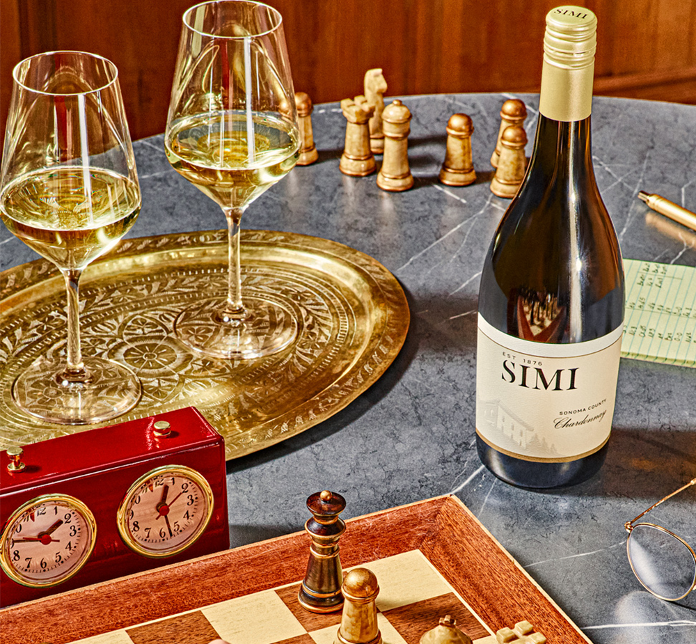 A bottle of SIMI Chardonnay sits on a chess table with two full wine glasses next to it.